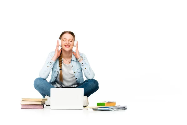 Chica feliz sentado en el suelo en los auriculares cerca de la computadora portátil, libros y copybooks aislados en blanco - foto de stock