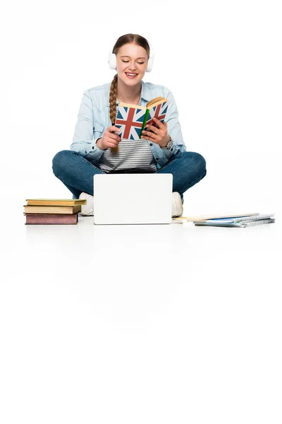 Lächelndes Mädchen, das Englisch auf dem Fußboden lernt, Kopfhörer in der Nähe von Laptop, Bücher und Textbücher isoliert auf weiß — Stockfoto