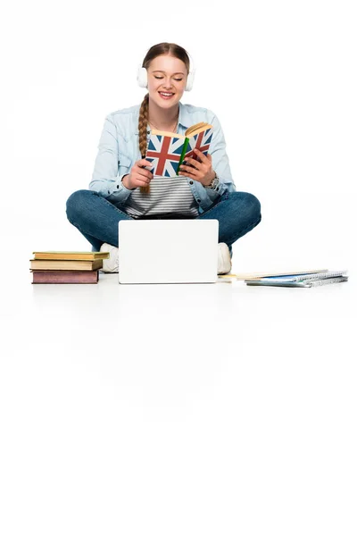 Niña sonriente aprendiendo inglés en el suelo en auriculares cerca de la computadora portátil, libros y copybook aislados en blanco - foto de stock