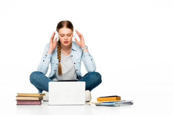 Fille assise sur le sol dans des écouteurs près d'un ordinateur portable, livres et copybooks isolés sur blanc — Photo de stock