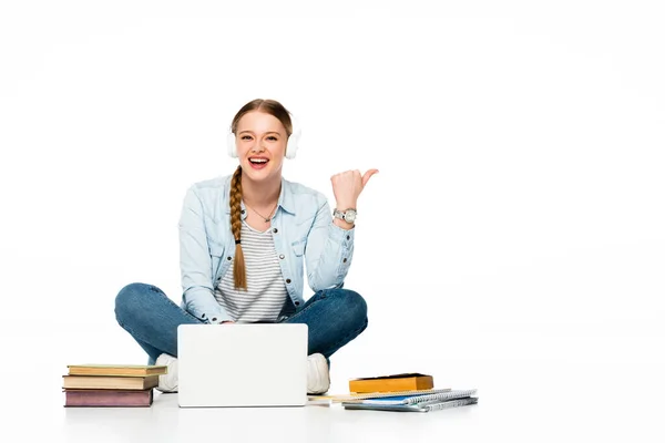 Fille souriante assise sur le sol dans des écouteurs près d'un ordinateur portable, des livres et des copybooks et pointant de côté isolé sur blanc — Photo de stock