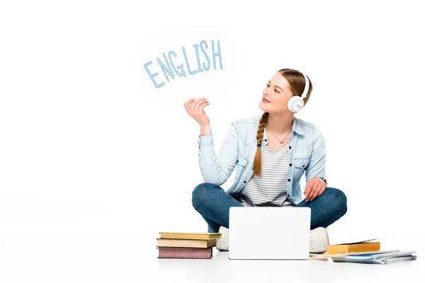 Fille souriante assise sur le sol dans des écouteurs avec bulle de parole avec lettrage anglais près d'un ordinateur portable, livres et copybooks isolés sur blanc — Photo de stock