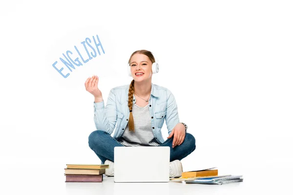 Smiling girl sitting on floor in headphones with speech bubble with english lettering near laptop, books and copybooks isolated on white — Stock Photo