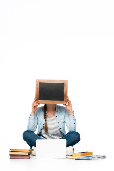 Ragazza seduta sul pavimento con viso oscuro vicino a laptop, libri e quaderni e tenendo la lavagna isolata su bianco — Foto stock