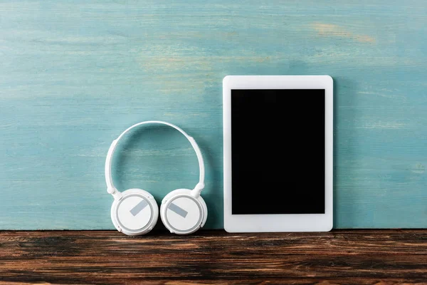 Auriculares, tableta digital con pantalla en blanco en la mesa de madera cerca de la pared azul - foto de stock