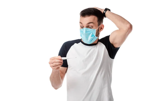 Шокированный человек в медицинской маске глядя на цифровой термометр изолирован на белом — стоковое фото