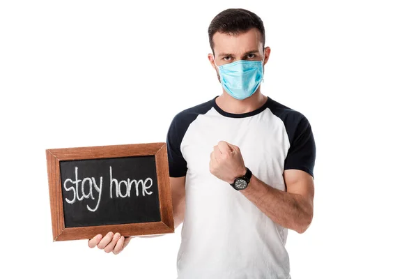 Homme dans le masque médical tenant tableau à craie avec rester à la maison lettrage et montrant poing isolé sur blanc — Photo de stock