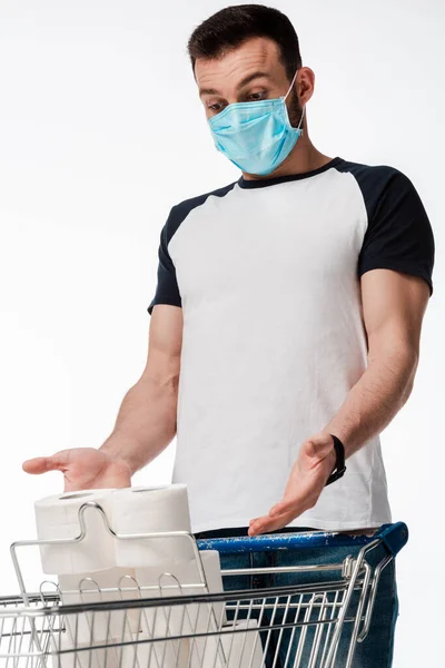 Man in medical mask gesturing while looking at shopping cart with toilet paper isolated on white — Stock Photo