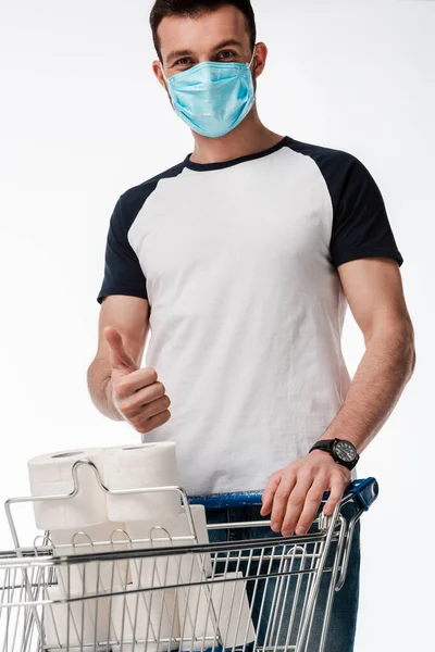 Uomo in maschera medica mostrando pollice verso l'alto vicino carrello della spesa con carta igienica isolato su bianco — Foto stock