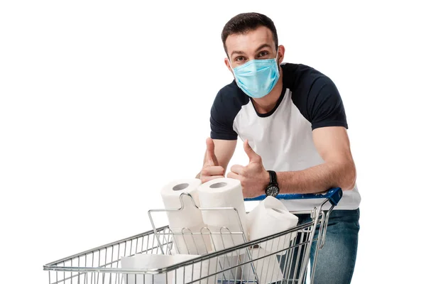 Мужчина в медицинской маске показывает большие пальцы вверх возле корзины с туалетной бумагой, изолированной на белом — стоковое фото
