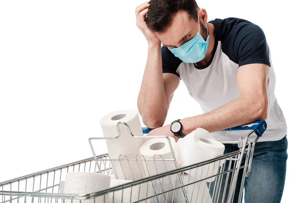 Uomo in maschera medica blu guardando carrello della spesa con carta igienica isolata su bianco — Foto stock