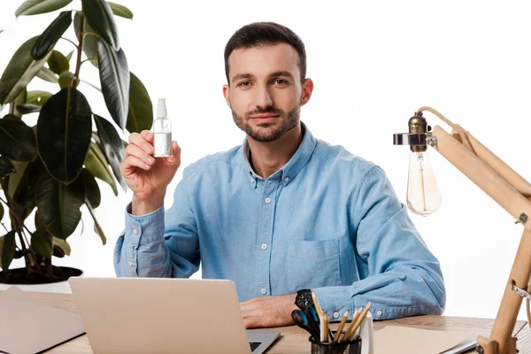 Guapo freelancer sosteniendo desinfectante de la mano cerca del ordenador portátil aislado en blanco - foto de stock