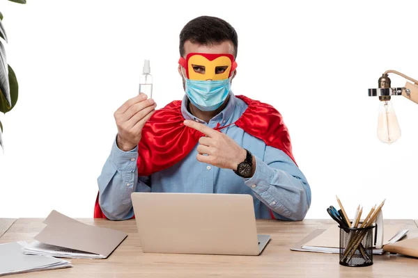 Homem em máscara médica e traje de super-herói apontando com o dedo na mão desinfetante perto laptop isolado no branco — Fotografia de Stock
