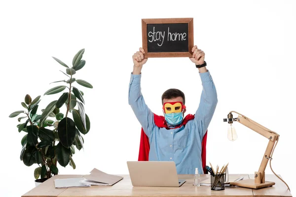 Man in medical mask and superhero cape holding chalk board with stay home lettering isolated on white — Stock Photo