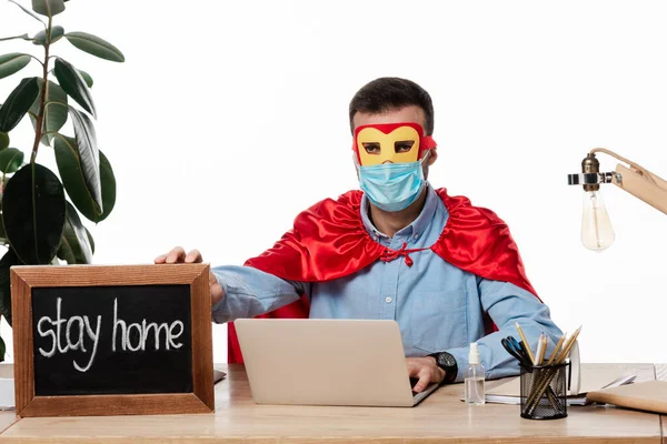 Freelancer em máscara médica e traje de super-herói segurando placa de giz com ficar em casa lettering isolado no branco — Fotografia de Stock