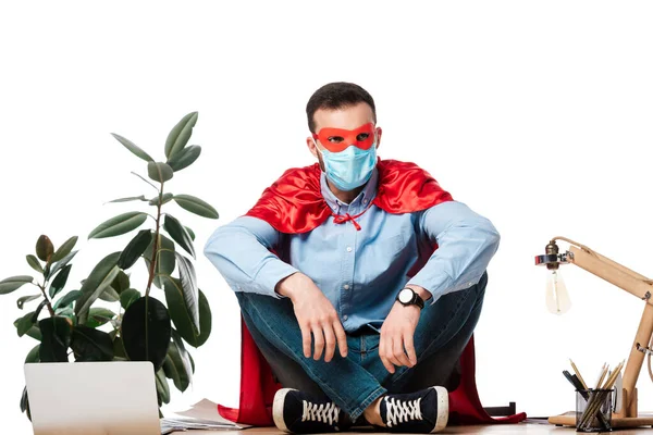 Homme en masque médical et costume de super-héros assis sur la table isolé sur blanc — Photo de stock