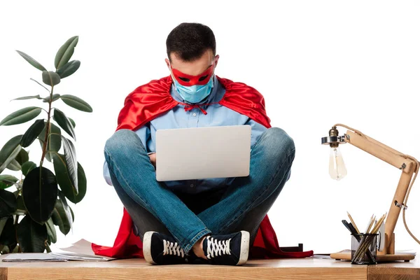 Uomo in maschera medica e mantello supereroe utilizzando il computer portatile e seduto sul tavolo isolato su bianco — Foto stock