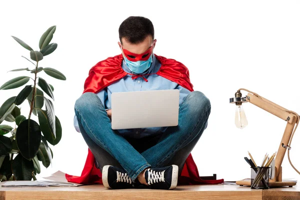 Hombre en máscara médica y capa de superhéroe usando el ordenador portátil y sentado en la mesa aislado en blanco - foto de stock