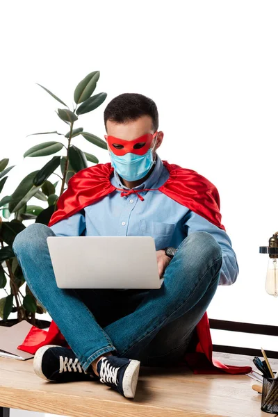 Freelance dans le masque médical et costume de super-héros en utilisant un ordinateur portable et assis sur la table isolé sur blanc — Photo de stock