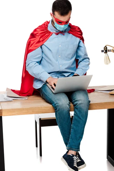 Freelancer in medical mask and superhero cape using laptop and sitting on table isolated on white — Stock Photo