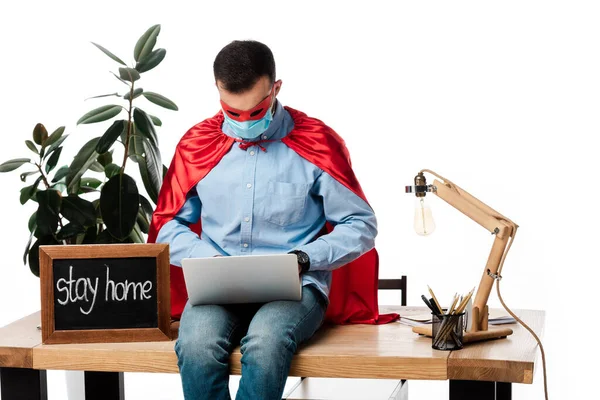 Фрилансер в медицинской маске и костюме супергероя, использующий ноутбук возле доски с надписью 