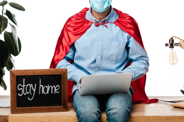 Visão cortada de freelancer em máscara médica e capa de super-herói usando laptop perto de placa de giz com ficar em casa lettering isolado em branco — Fotografia de Stock