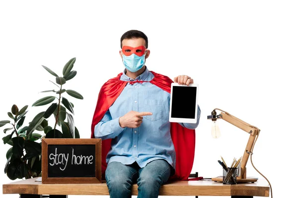 Hombre en máscara médica y capa de superhéroe apuntando con el dedo a la tableta digital con pantalla en blanco cerca de pizarra con letras estancia en casa aislado en blanco - foto de stock