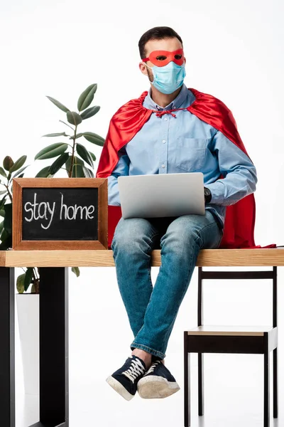 Freelancer em máscara médica e capa de super-herói usando laptop perto de placa de giz com ficar em casa lettering isolado no branco — Fotografia de Stock