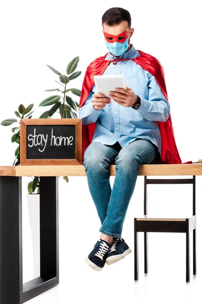 Hombre en máscara médica y capa de superhéroe utilizando tableta digital cerca de pizarra con letras estancia en casa aislado en blanco - foto de stock