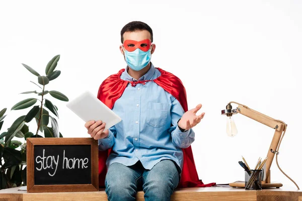 Homem em máscara médica e capa de super-herói segurando tablet digital perto de placa de giz com ficar em casa letras isoladas em branco — Fotografia de Stock