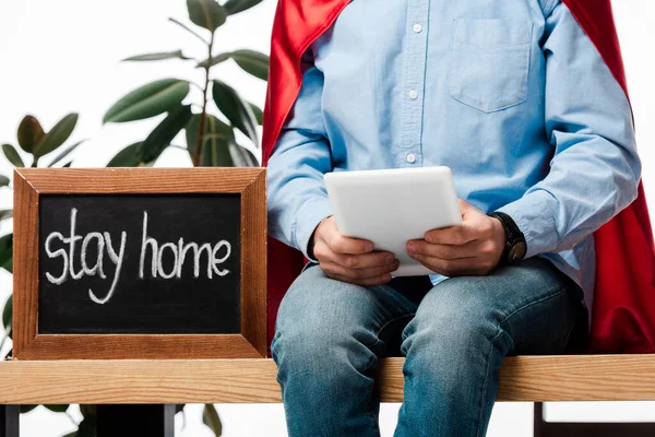 Cropped view of man in superhero cape using digital tablet near chalk board with stay home lettering isolated on white — Stock Photo