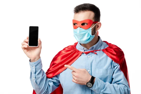 Homem em traje de super-herói e máscara médica apontando com o dedo para o smartphone com tela em branco isolado no branco — Fotografia de Stock
