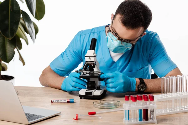 Científico en máscara médica y gafas tocando microscopio cerca de tubos de ensayo con letras coronavirus y portátil aislado en blanco - foto de stock