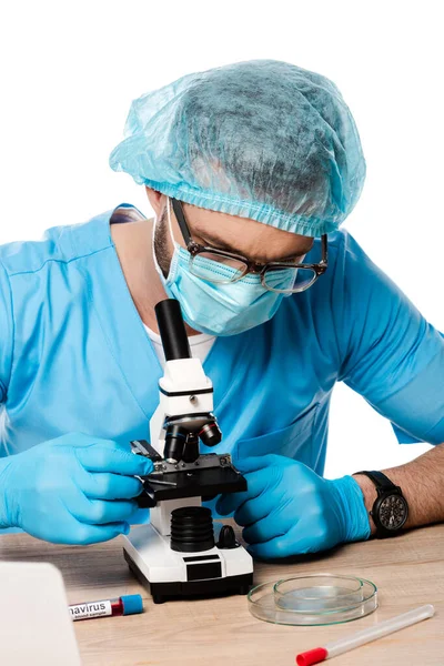 Scientist in medical cap and glasses touching microscope near test tubes with coronavirus lettering isolated on white — Stock Photo