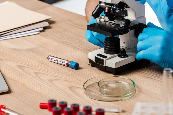 Vista recortada del científico en guantes de látex tocando el microscopio cerca de tubos de ensayo con letras de coronavirus en la mesa - foto de stock