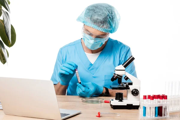 Scientist in medical mask and goggles holding test tube with coronavirus lettering near microscope and laptop isolated on white — Stock Photo