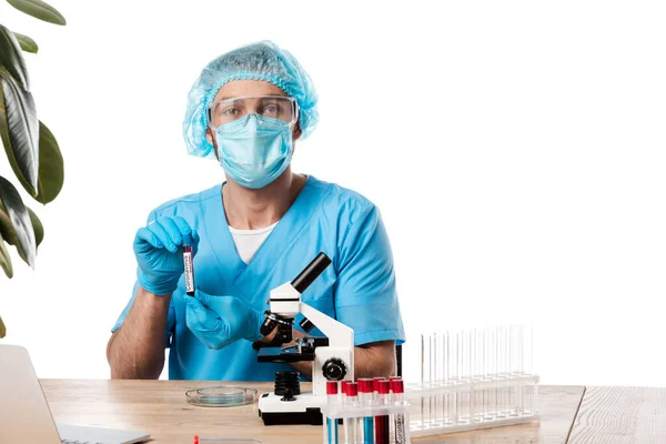 Científico en gorra médica y gafas que sostienen el tubo de ensayo con letras de coronavirus cerca del microscopio y el ordenador portátil aislado en blanco - foto de stock