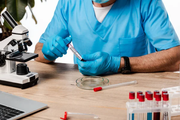 Vista ritagliata dello scienziato in guanti di lattice in possesso di provetta con coronavirus lettering vicino al microscopio — Foto stock