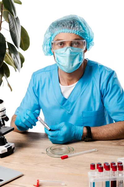 Вчений у медичній шапці та окулярах, що тримає пробірку з коронавірусною плямою біля мікроскопа ізольовано на білому — стокове фото