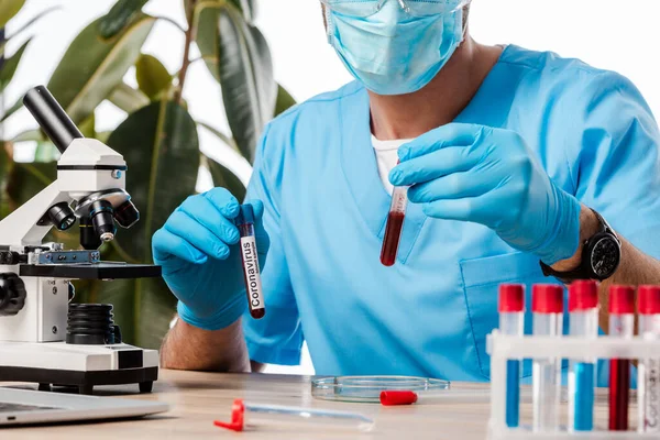 Ausgeschnittene Ansicht eines Wissenschaftlers im Latexhandschuh, der Reagenzgläser mit Blutproben und Coronavirus-Schriftzug in der Nähe des Mikroskops hält — Stockfoto