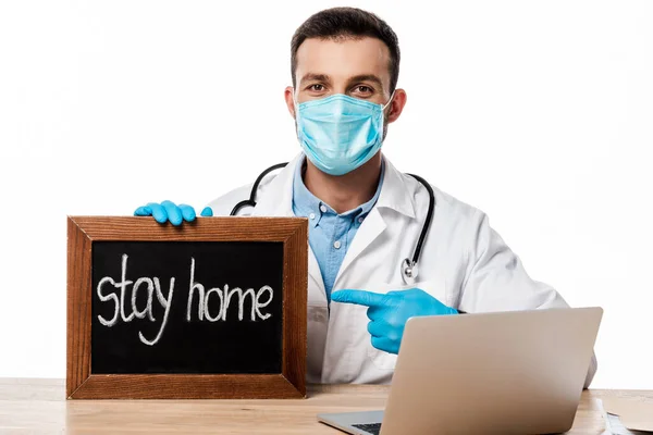 Médico en máscara médica y abrigo blanco apuntando con el dedo a la pizarra con letras de la estancia en casa aislado en blanco - foto de stock