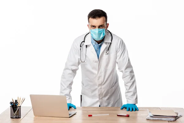 Doctor in medical mask and white coat standing near laptop and test tubes isolated on white — Stock Photo