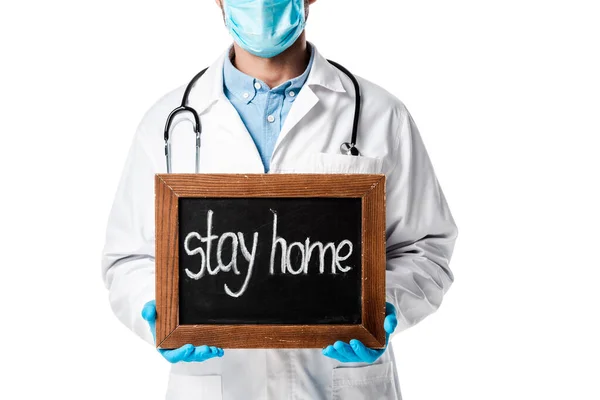 Vista ritagliata del medico in maschera medica e bianco cappotto tenendo bordo gesso con soggiorno casa lettering isolato su bianco — Foto stock