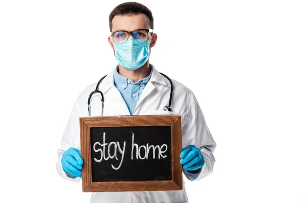 Médico en máscara médica, gafas y abrigo blanco sosteniendo pizarra con letras de estancia en casa aislado en blanco - foto de stock