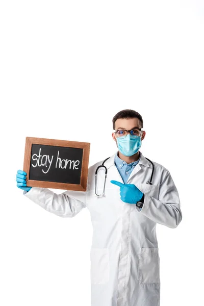 Medico in maschera medica e cappotto bianco che punta con il dito a bordo gesso con soggiorno casa lettering isolato su bianco — Foto stock