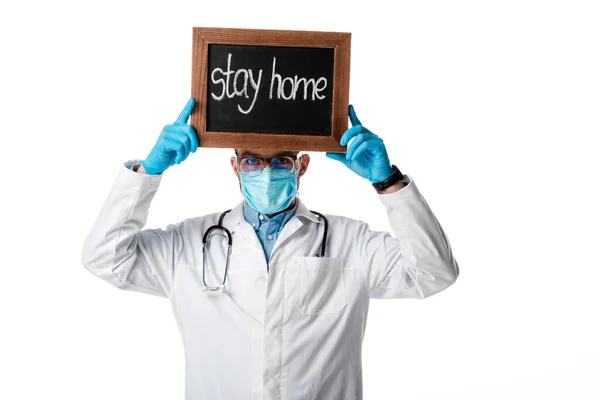 Medico in maschera medica e bianco cappotto tenendo bordo gesso con soggiorno casa lettering sopra la testa isolato su bianco — Foto stock