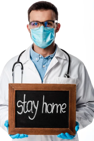 Medico in maschera medica e bianco cappotto tenendo bordo gesso con soggiorno casa lettering isolato su bianco — Foto stock