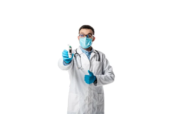 Médico en máscara médica y abrigo blanco sosteniendo pirómetro y mostrando el pulgar hacia arriba aislado en blanco — Stock Photo