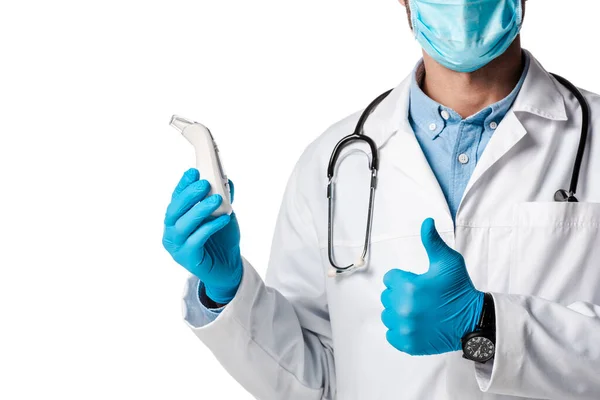 Ausgeschnittene Ansicht des Arztes in medizinischer Maske und weißem Kittel, der ein berührungsloses Pyrometer hält und den Daumen isoliert auf weiß zeigt — Stockfoto
