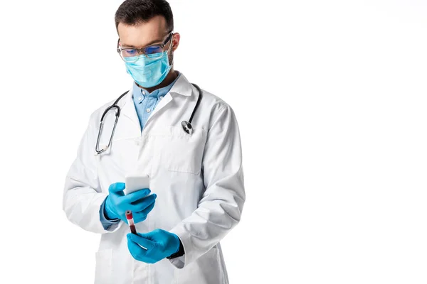 Médecin en masque médical et manteau blanc tenant smartphone et prenant une photo du tube à essai avec échantillon de sang isolé sur blanc — Photo de stock